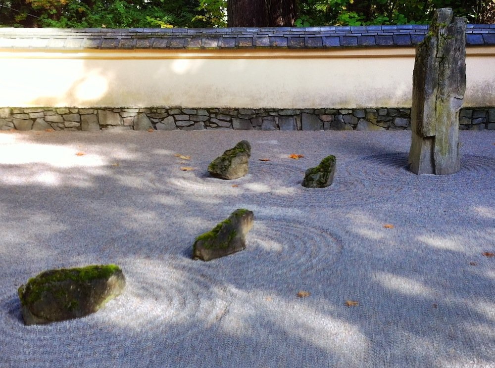 A pebbled Zen garden with scattered large rocks with ripples drawn in the pebbles around them. The back wall is in the Japanese style. Above, the outlines of a Pacific Northwest forest can be seen