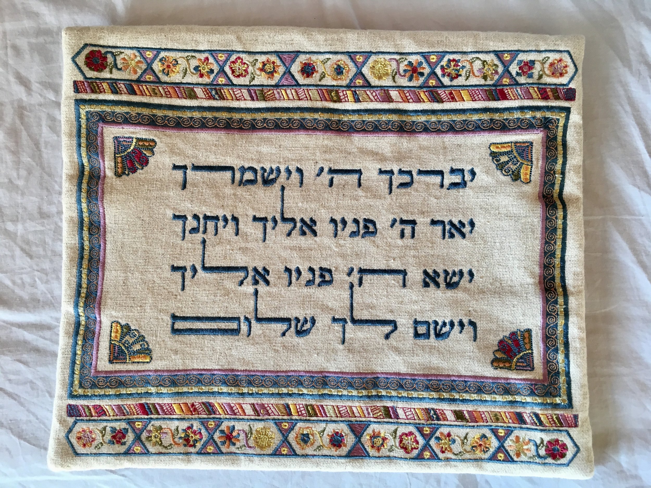A white tallit bag embroidered with flowers and colorful borders bearing the text of the priestly blessing in Hebrew.;
