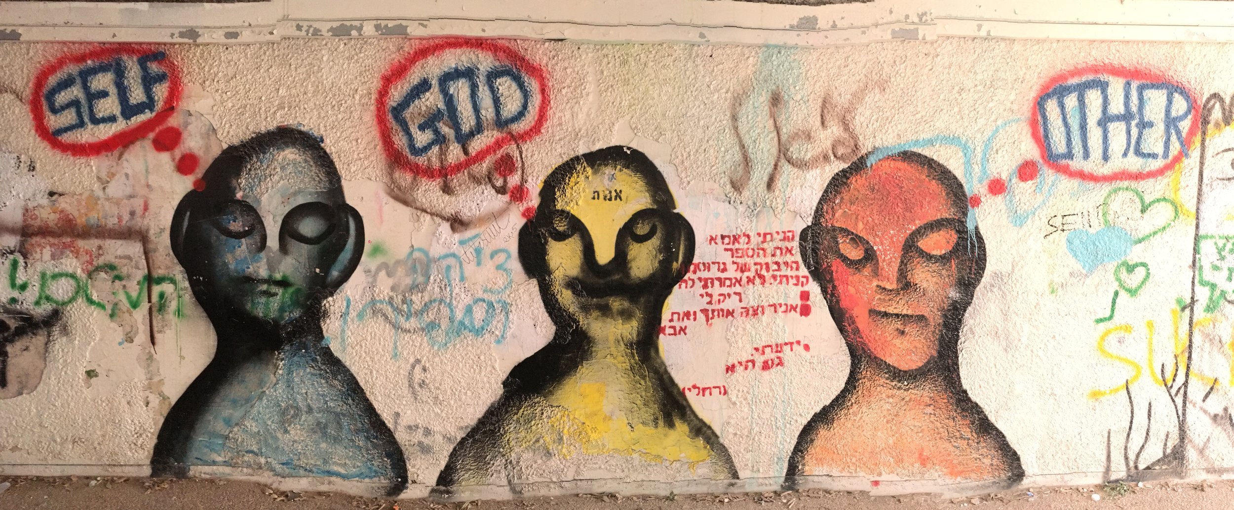 A spray painted mural inside a pedestrian tunnel with three humanoid figures, one blue, one yellow, and one orange, with thought bubbles above their heads reading "Self," "God," and "Other." The forehead of the yellow one ("God") says "emet" (truth)