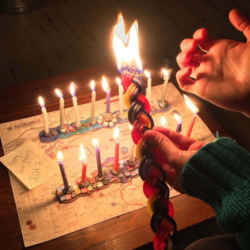 Two makeshift hanukiot with all the candles lit, a woman's hands holding a lit braided havdalah candle, and a note on the table next to the candles that reads "Happy New Year! 2017"