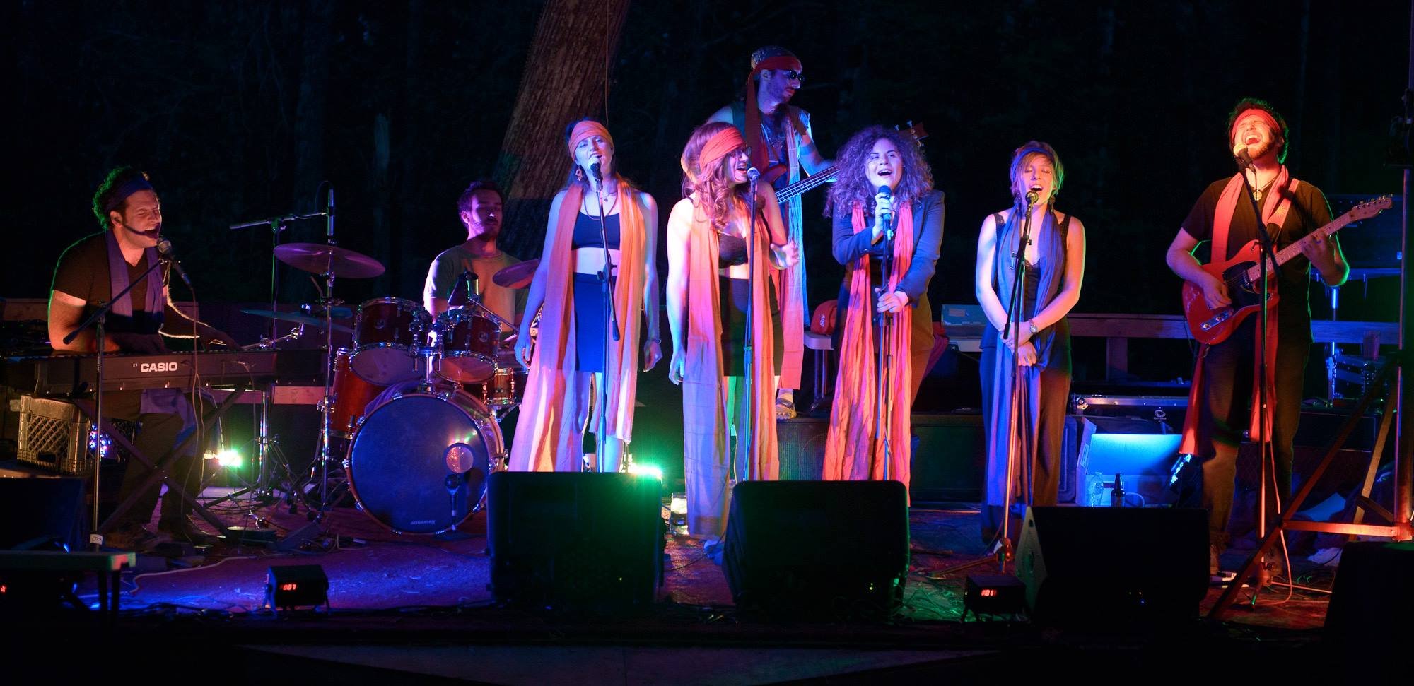 An eight-person rock band (keys, drums, bass, guitar, four vocalists) dressed in black with long red headscarves playing on stage at night under blue and green lights. The trunk of a tree is visible behind the stage.