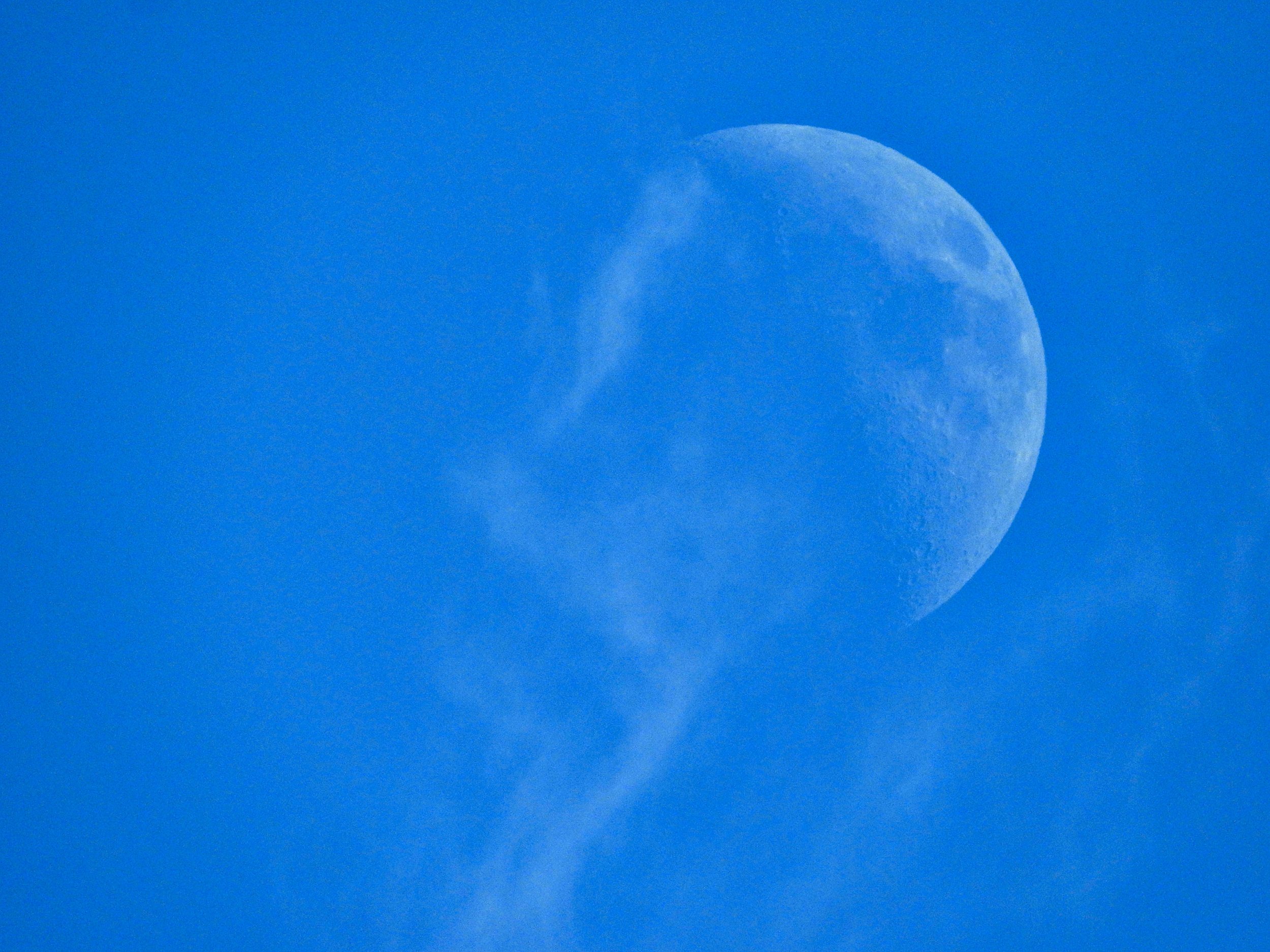A waxing crescent Moon before the first quarter in the afternoon sky with wisps of clouds looking like they're dissolving off of it.