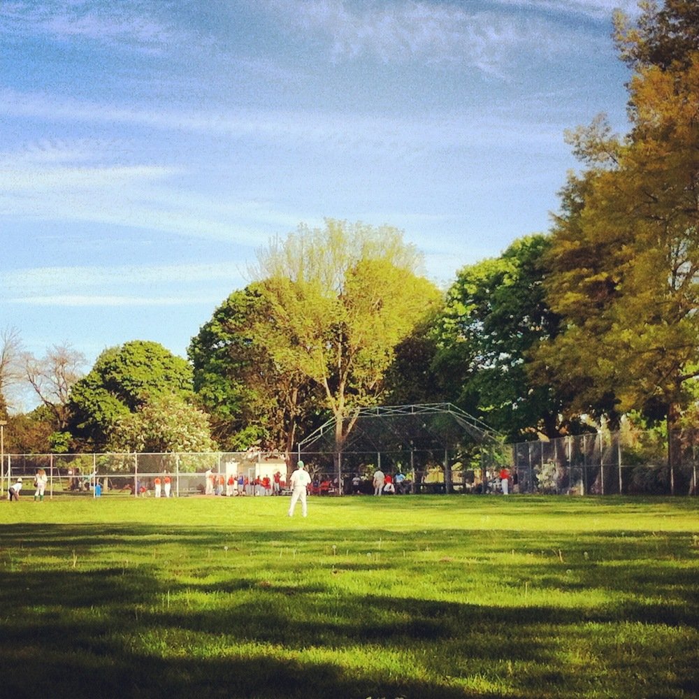 A heavily filtered square photo of a baseball field where a youth game is taking place, taken from the grass beyond left field